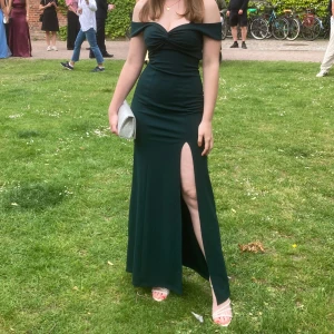 Mörkgrön långklänning med slits - Elegant mörkgrön långklänning med offshoulder-design och en slits framtill. Perfekt för festliga tillfällen. Klänningen har en figurnära passform som framhäver silhuetten. Endast använd en gång på min studentbal köpt för 1200kr