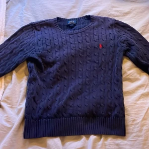 Mörkblå stickad tröja från Polo Ralph Lauren - Säljer en snygg mörkblå stickad tröja från Polo Ralph Lauren. Den har ett klassiskt kabelstickat mönster och en liten röd logga på bröstet. Perfekt för höst och vinter, med ribbade muddar vid ärmslut och nederkant. Passar både till vardags och lite finare tillfällen. Storlek L i barn den är ytterst liten för killar som har s i män. Kan gå ner i pris vid snabb affär.