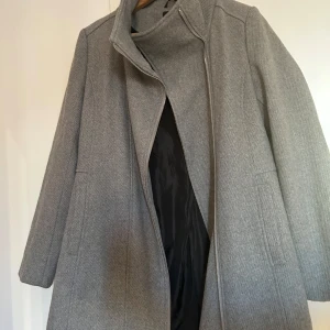 Grå kappa med dragkedja - Säljer en stilren grå kappa i mycket bra skick. Den har en hög krage och stängs med dragkedja. Perfekt för höst och vinter, och ger en elegant look. Passar både till vardags och lite finare tillfällen.