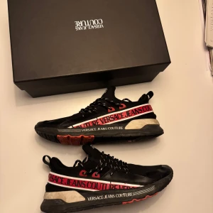 Svarta sneakers från Versace Jeans Couture - Säljer ett par snygga svarta sneakers från Versace Jeans Couture. Skorna har en cool design med röd och vit text längs sidan och svarta snören. Perfekta för både vardag och fest! De är i okej skick men har mycket kvar att ge.och kommer i originalkartong.