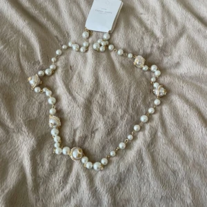 Pärlhalsband med gulddetaljer - Elegant långt pärlhalsband med vita pärlor och gulddetaljer. Perfekt för att ge en touch av glamour till din outfit. Halsbandet har en klassisk design med större dekorerade pärlor som ger det en unik stil. Passar både till vardags och festliga tillfällen.
