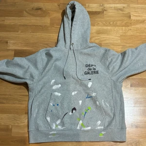 Grå hoodie från GALLERY DEPT - Säljer en grå hoodie från gallery dept i storlek xl. med coola färgstänk i vitt, blått, grönt och svart. Den har en stor ficka fram och justerbar huva. Perfekt för en avslappnad stil eller konstnärlig look. Passar både till vardags och chillkvällar.Tar även byten