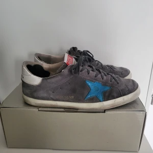 Grå sneakers från Golden Goose - Säljer ett par grå sneakers från Golden Goose i bra skick. De har en cool blå stjärna på sidan och vit häl med märkesnamnet. Skorna har snörning och är gjorda i mocka. Perfekta för en avslappnad stil med en touch av lyx. Kommer med originalförpackning.