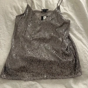 Glittrig topp från New Yorker - Säljer en superfin glittrig topp från New Yorker. Den har smala axelband och en v-ringad design. Perfekt för festliga tillfällen. Toppen är i nytt skick med prislappen kvar.