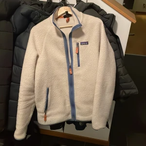 Beige fleecejacka från Patagonia - Säljer en supermysig vit fleecejacka från Patagonia i storlek M. Den har en snygg blå dragkedja och praktiska fickor med dragkedjor. Perfekt för kyliga höst- och vinterdagar. Jackan är i mycket bra skick och redo för nya äventyr!