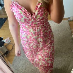 Blommig sommarklänning med volangdetaljer - Söt blommig klänning i rosa och grönt med volangdetaljer. Perfekt för sommarens fester eller en dag på stan. Klänningen har en smickrande passform och ärmlös design, vilket gör den sval och bekväm. Ett måste för garderoben!