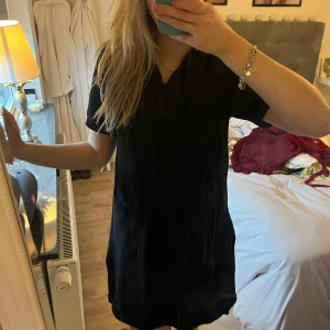 Svart kortklänning - Säljer en stilren svart kortklänning med korta ärmar och praktiska fickor framtill. Perfekt för en enkel och chic look. Klänningen har en avslappnad passform och är lätt att matcha med olika accessoarer. Stickad