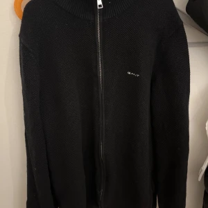 full zip gant - Säljer en stilren svart stickad tröja från GANT med dragkedja framtill. Tröjan har långa ärmar och en diskret logga på bröstet. Perfekt för kyliga dagar.