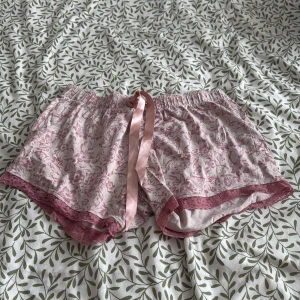 Rosa blommiga shorts med spetsdetaljer - Säljer ett par söta rosa shorts med blommigt mönster och spetsdetaljer. De har en bekväm elastisk midja och en dekorativ rosett framtill. Perfekta för en avslappnad dag hemma eller sommarens varma dagar.