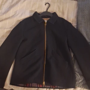 Svart jacka från NN07 - Säljer en stilren marinblå jacka från NN07 med dragkedja framtill. Jackan har en klassisk krage och två snedställda fickor. Insidan är fodrad med ett rutigt mönster. Perfekt för en elegant look.