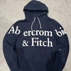 Mörkblå hoodie från Abercrombie & Fitch - Säljer en snygg mörkblå hoodie från Abercrombie & Fitch med stor vit logga på framsidan. Tröjan har en klassik look med storlek medium, passar även Large. Skicket är 9/10. Perfekt för en vardaglig Hoodie. Hör av vid funderingar. 😁