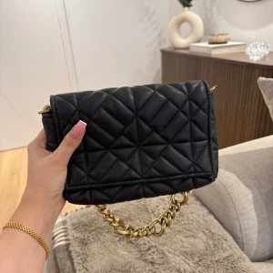 Svart quiltad axelväska med kedja - Snygg svart quiltad axelväska från Zara med en iögonfallande guldkedja som axelrem. Perfekt för att ge din outfit en lyxig touch. Väskan har en praktisk storlek och ett stilrent mönster.