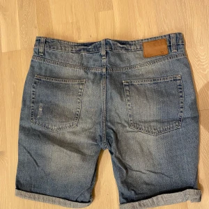 Blå jeansshorts från 157 - Snygga blå jeansshorts från 157 storlek Large med slitna detaljer och uppvikta ben. De har en klassisk femficksdesign och stängs med knapp och dragkedja. Perfekta för en avslappnad stil. Använda men bra skick.