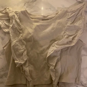 Beige ribbad topp från Zara - Säljer en söt beige ribbad topp från Zara med volangdetaljer längs ärmarna. Toppen är ärmlös och har en rund halsringning. Säljer efter som att den är för liten.