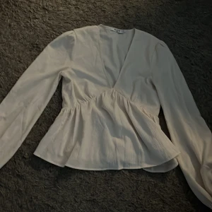 Vit blus från NA-KD - Säljer en stilren vit blus från NA-KD med v-ringning och långa ärmar. Blusen har en lätt rynkad midja som ger en fin passform. Perfekt för att skapa en elegant look.