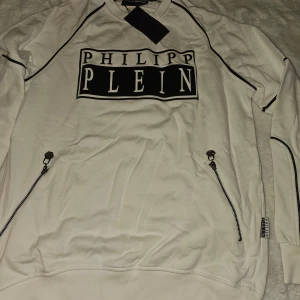 Vit tröja från Philipp Plein - Snygg vit tröja från Philipp Plein med svart logotyp på bröstet. Tröjan har långa ärmar och dragkedjor på sidorna för en cool look. Perfekt för en stilren och modern outfit.