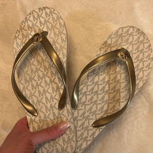 Michael Kors tofflor - Snygga flip-flops från Michael Kors med guldiga remmar och logotypdetalj. Sulan har ett diskret mönster i beige och vitt. Perfekta för sommardagar på stranden eller i stan.