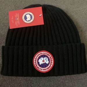 Svart ribbad mössa från Canada Goose - Snygg svart ribbad mössa från Canada Goose med ett broderat märke framtill. Perfekt för kyliga dagar och ger en stilren look. Mössan är i en klassisk design som passar de flesta.vill ni ha fler bilder är de bara att fråga! Priset går att pruta!👍