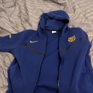 Blå hoodie med dragkedja och logga Niketec - Säljer en blå hoodie från Nike med dragkedja och huva. Den har en gul logga på bröstet och ärmen. Perfekt för sportiga tillfällen eller en avslappnad stil.