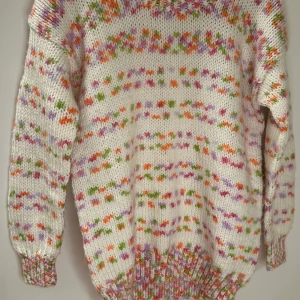 Flerfärgad stickad tröja - Säljer en mysig handstickad tröja i vitt med flerfärgat mönster i rosa, orange och grönt. Tröjan har långa ärmar och ribbstickade muddar vid ärmslut och nederkant. Perfekt för kyliga dagar!