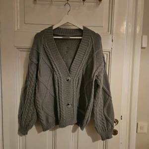 Grå stickad kofta från The Tortured Poets Department - OFFICIELL TAYLOR MERCH. Säljer en mysig grå stickad kofta från The Tortured Poets Department. Koftan har en djup v-ringning och stora knappar framtill. Perfekt för kyliga dagar med sitt snygga kabelstickade mönster.