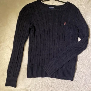 Mörkblå stickad tröja från Ralph Lauren - Säljer en stilren mörkblå stickad tröja från Ralph Lauren med kabelstickat mönster. Tröjan har långa ärmar och en liten rosa logga på bröstet. Perfekt för kyligare dagar.🩷🩷