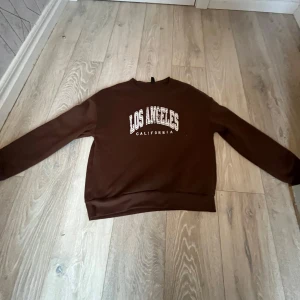 Brun sweatshirt med Los Angeles-tryck - Säljer en brun sweatshirt med texten 'Los Angeles California' i vitt tryck på framsidan. Tröjan har långa ärmar och en klassisk rund halsringning. Perfekt för en avslappnad stil.