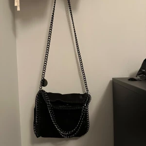 Svart axelväska med kedjedetaljer - Snygg svart axelväska med kedjedetaljer och magnetknapp. Väskan har en stilren design med en kedja som axelrem och flera fack inuti för enkel organisering. Perfekt för att ge en edgy touch till din outfit. Fint skick och den har inga skador. Använt fåtal gånger. 