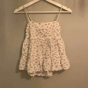 Blommig linne från Gina Tricot - Söt blommig linne från Gina Tricot i vitt med små blommor i rosa och blått. Toppen har smala axelband och en volangdetalj nedtill. Den har en öppen rygg med knytband för en justerbar passform. Banden har jag förkortat själv och sytt fast igen.💕