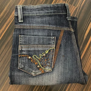 Lågmidjade bootcut jeans med detaljer - Lågmidjade jeans med fina broderade detaljer. Byxorna är även utsvängda nertill. Säljer då de inte passar mig längre. Finns en defekt (se sista bilden). Pris kan diskuteras och kan gå ner i pris 🩷🩷