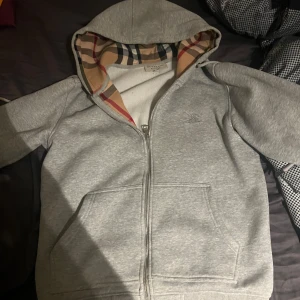 Grå huvtröja från Burberry - Säljer en stilren grå huvtröja från Burberry med klassiskt rutigt mönster i huvan. Tröjan har dragkedja framtill och en broderad logga på bröstet. Perfekt för en avslappnad look.