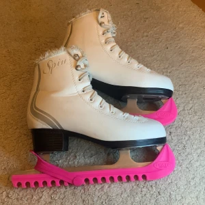 Vita konståkningsskridskor med rosa skydd - Säljer ett par vita konståkningsskridskor från Spin med fluffig kant och snörning. De har rosa skydd på skenorna och en klassisk design med svart klack. Perfekta för vinterns isäventyr!