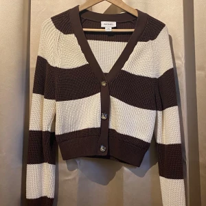 Brun och vit randig kofta från Monki - Säljer en snygg brun och vit randig kofta från Monki. Den är stickad med långa ärmar och har knappar framtill. Perfekt för en stilren look!