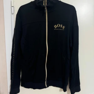 Hugo Boss tröja S - Snygg svart jacka från Hugo Boss med dragkedja framtill och logotyp på bröstet. Jackan har långa ärmar och en stilren design som passar perfekt för en casual look.