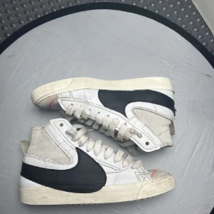 Nike Blazer Mid sneakers i vitt och svart - Snygga Nike Blazer Mid sneakers i vitt med svart swoosh. Skorna har en klassisk hög design med snörning och en robust sula. Perfekta för en stilren look.