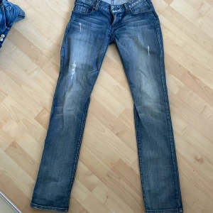 Blå jeans från Guess - Snygga blå jeans från Guess med en klassisk femficksdesign. De har en straight passform och är dekorerade med subtila slitningar för en trendig look. Dem är lite sydda vid låren för att få dem lite tightare i rumpan