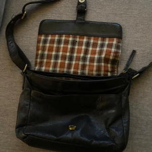 Svart axelväska med rutigt foder - Snygg svart axelväska i skinn med ett klassiskt rutigt foder i brunt, beige och vitt. Väskan har en justerbar axelrem och stängs med ett spänne. Perfekt för att bära dina dagliga nödvändigheter med stil. Vintage 