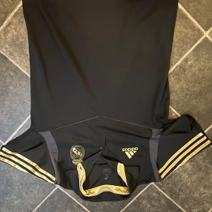 Svart fotbollströja från real madrid - Snygg svart fotbollströja från real Madrid med guldiga detaljer och klubbmärke på bröstet. Tröjan har korta ärmar med guldiga ränder och en stilren design. 