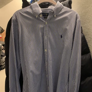 Randig skjorta från Ralph Lauren - Snygg blå- och vitrandig skjorta från Ralph Lauren i slim fit. Skjortan är tillverkad i bomull med stretch och har en broderad logotyp på bröstet. Perfekt för en stilren look.ny pris 1800kr mitt pris 499kr