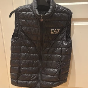 Svart dunväst från EA7 - Snygg svart dunväst från EA7 med dragkedja framtill. Västen har en quiltad design och är perfekt för kyligare dagar. Den har en stilren look med EA7-loggan på bröstet.