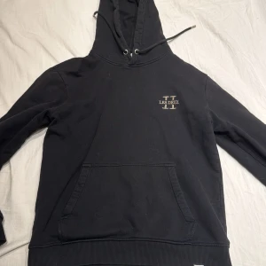 Svart hoodie från Les Deux - Säljer en stilren svart hoodie från Les Deux med en praktisk magficka och justerbar huva. Perfekt för en avslappnad look. Diskret logga på bröstet.