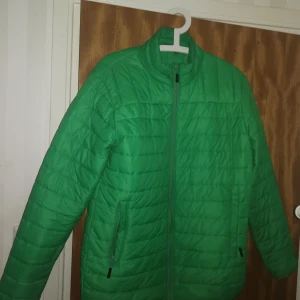 Grön pufferjacka - Snygg grön pufferjacka med dragkedja och hög krage. Jackan har en quiltad design och praktiska fickor med dragkedjor. Perfekt för kyligare dagar.