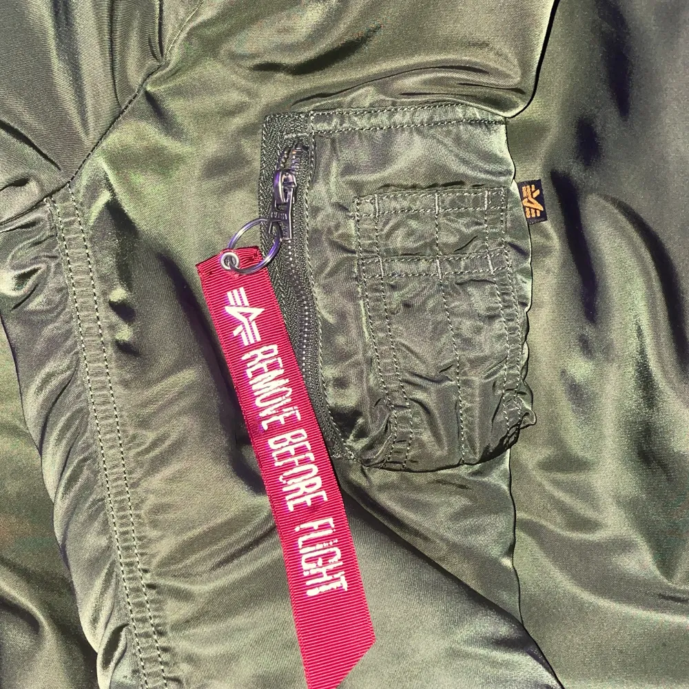 Snygg grön bomberjacka från Alpha Industries med orange foder och avtagbar 'Remove Before Flight'-tag. Jackan har dragkedja och en mysig huva med fuskpäls. Perfekt för kyligare dagar.. Takit.