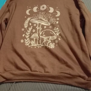 Brun tröja med svampmotiv - Säljer en brun tröja med ett unikt tryck av svampar och månfaser. Tröjan har långa ärmar och är perfekt för en avslappnad stil. Passar bra till höst och vår.