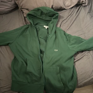 Grön hoodie från Lacoste - Säljer en grön hoodie från Lacoste med dragkedja och klassisk passform. Den har en broderad logga på bröstet och praktiska fickor framtill. Perfekt för en avslappnad stil.