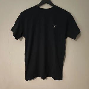 Svart t-shirt från Gant - Snygg svart t-shirt från Gant med liten logga på bröstet. Perfekt för en stilren och enkel look. Tillverkad i mjukt material för bekväm passform. Den är i storlek för 158/164. Den är väldigt lite använd. 