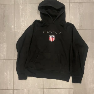 Svart hoodie från GANT - Säljer en svart hoodie från GANT med en broderad logga och ett emblem på framsidan. Tröjan har en klassisk känguruficka och en bekväm huva. Perfekt för en avslappnad stil.