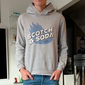 Grå hoodie från Scotch & Soda - Snygg grå hoodie från Scotch & Soda med ett stort tryck i blått och vitt på framsidan. Perfekt för en avslappnad stil. Tröjan har en riktigt snygg passform. Storleken är 16 år modellen på bilden är 170 cm