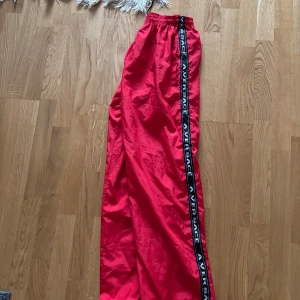 Röda byxor från Aversace - Snygga röda byxor från Aversace med elastisk midja och svarta sidoremsor med logotyp. Byxorna har en broderad logga på baksidan och är perfekta för en avslappnad stil.