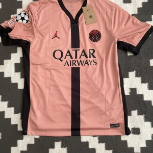 Rosa PSG fotbollströja med Mbappé 7 - Snygg rosa PSG-tröja från Nike med Mbappé 7 på ryggen. Tröjan har korta ärmar och svarta detaljer, inklusive klubbmärke och sponsorlogga. Perfekt för fotbollsfans som vill visa sitt stöd för laget och spelaren.